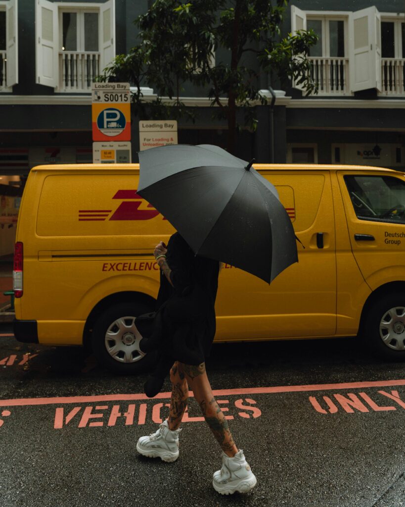 A woman with an umbrella walks past a yellow delivery van on a rainy street in Singapore.